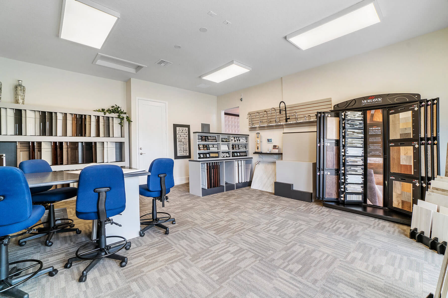 The interior of this Akel Homes showroom features blue chairs around a table, display racks with various flooring samples, and organized shelving with samples adorning the walls.