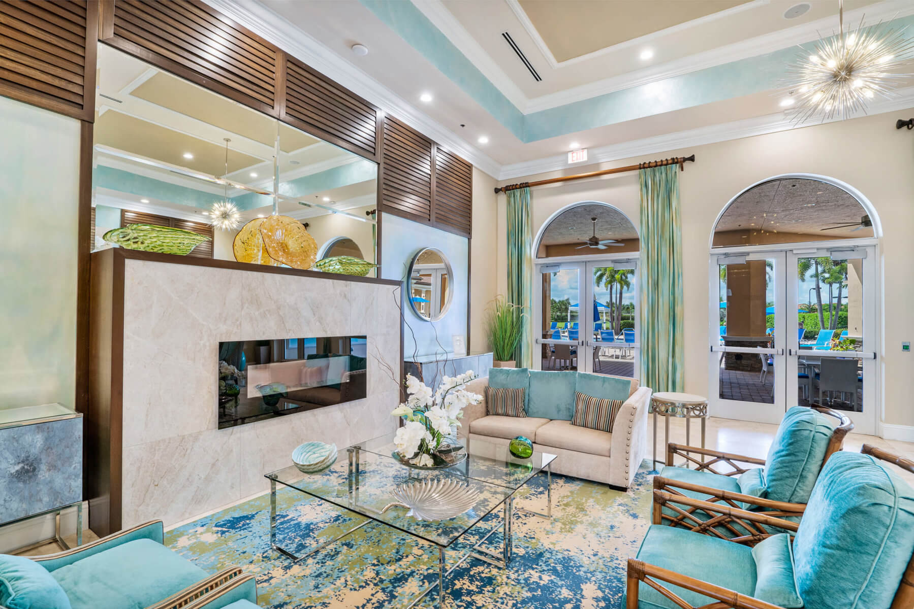 A modern living room by Akel Homes features a decorative ceiling, large mirror, and glass coffee table. The space includes a comfortable sofa, stylish armchairs, and decorative pillows. Elegant curtains frame the large arched windows, completing the sophisticated ambiance.