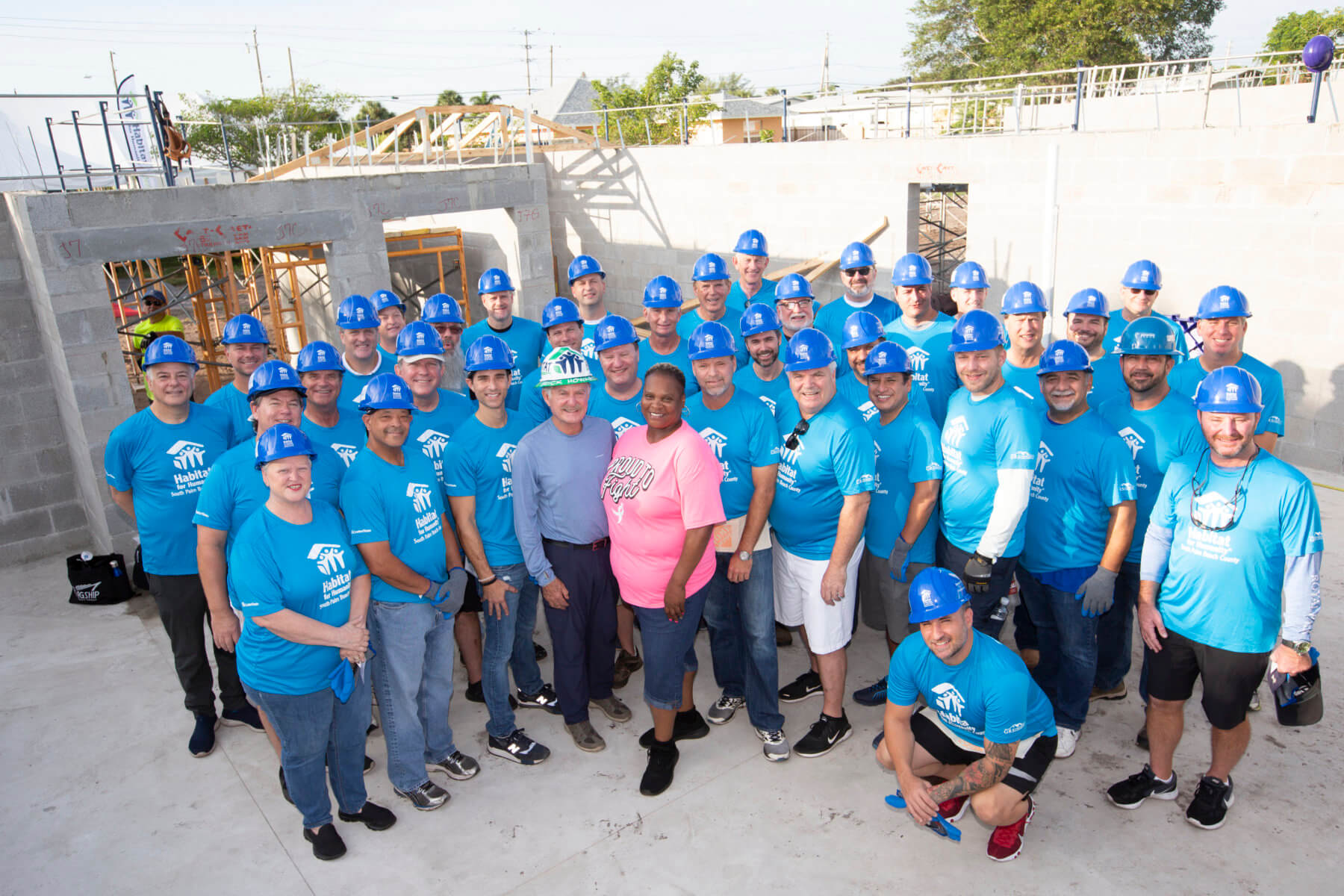 A group of people wearing blue shirts and blue hard hats stands together at an Akel Homes construction site.