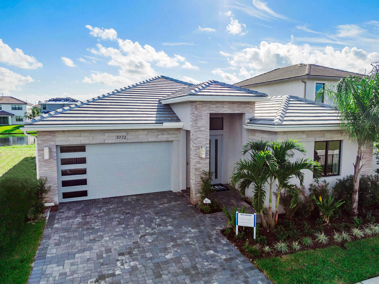 Single-story modern house by Akel Homes features a tiled roof and lush landscaping. A brick driveway guides you to the garage, adorned with frosted windows. The scene is set against a bright blue sky with scattered clouds, illustrating the perfect blend of style and comfort.