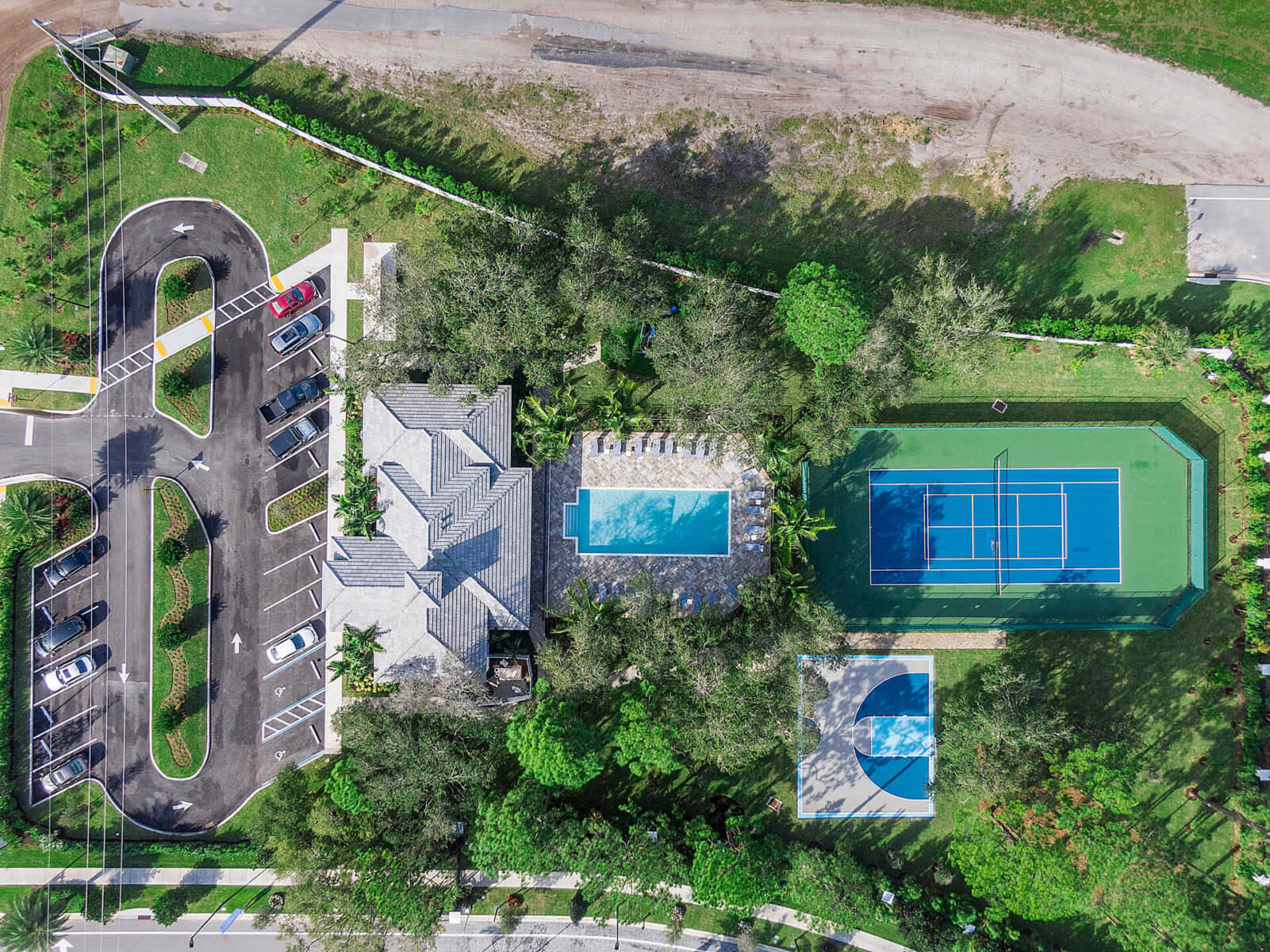 Aerial view of a recreational area by Akel Homes featuring a parking lot, a building with a pool, a tennis court, and a basketball court nestled among lush trees.