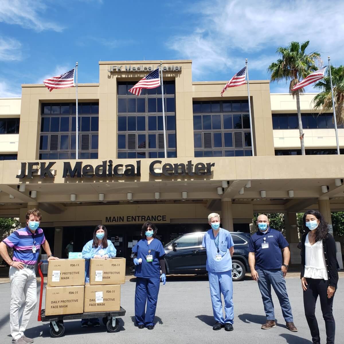 A group of people wearing masks stands in front of JFK Medical Center's main entrance, with boxes labeled "face masks" on a cart donated by Akel Homes. Flags are visible above the building.