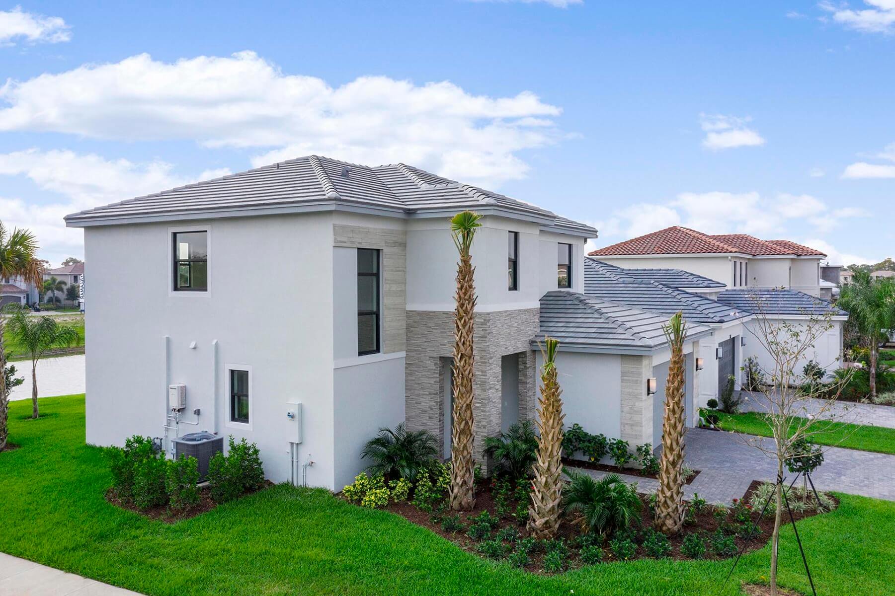 A two-story white house crafted by Akel Homes, featuring a tile roof, is nestled among palm trees and a well-maintained lawn beneath a blue sky dotted with clouds.