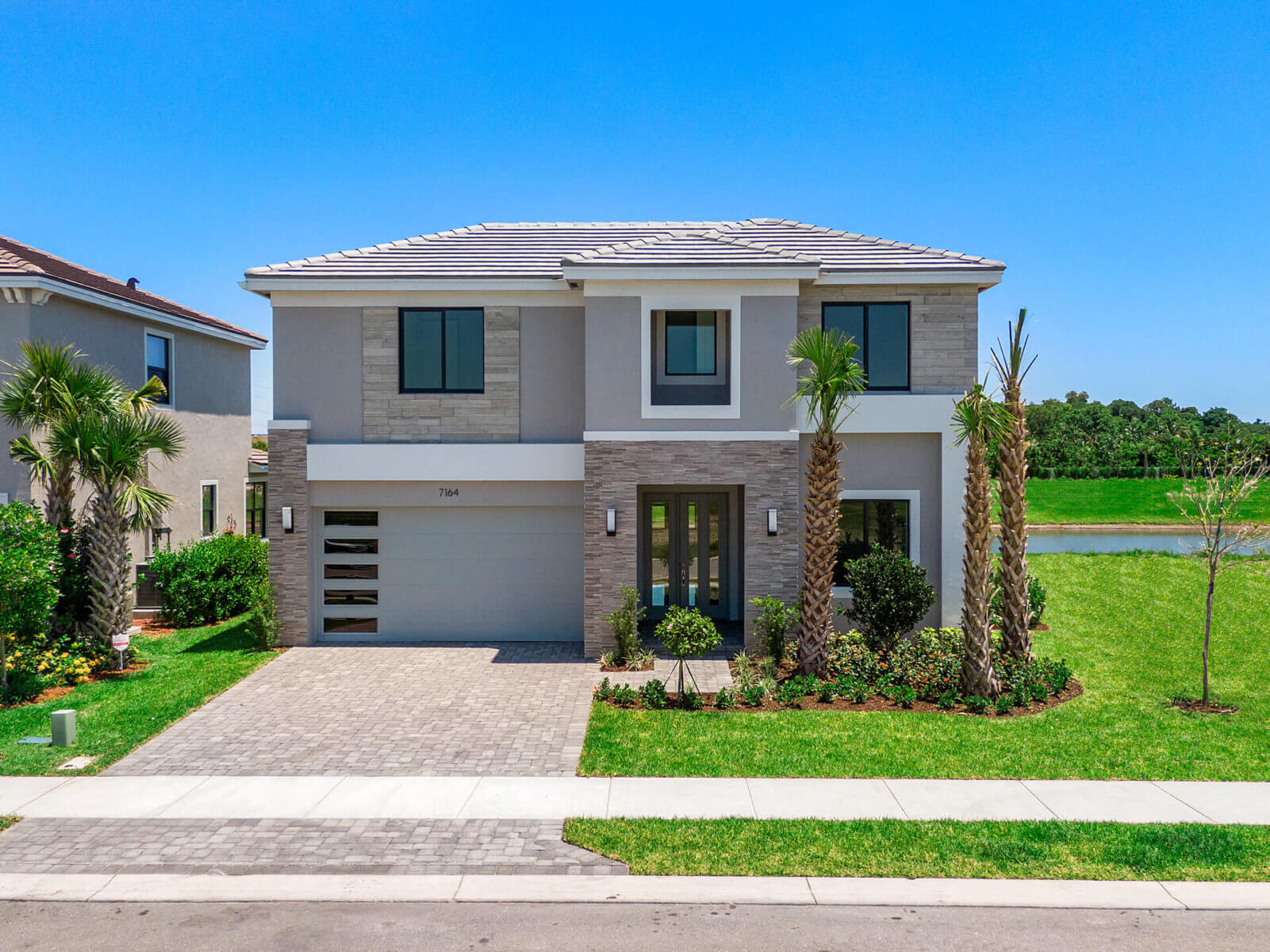 A modern two-story Akel Homes house boasts a stone facade, two-car garage, and landscaped yard, all set against the serene backdrop of a blue sky and shimmering lake.