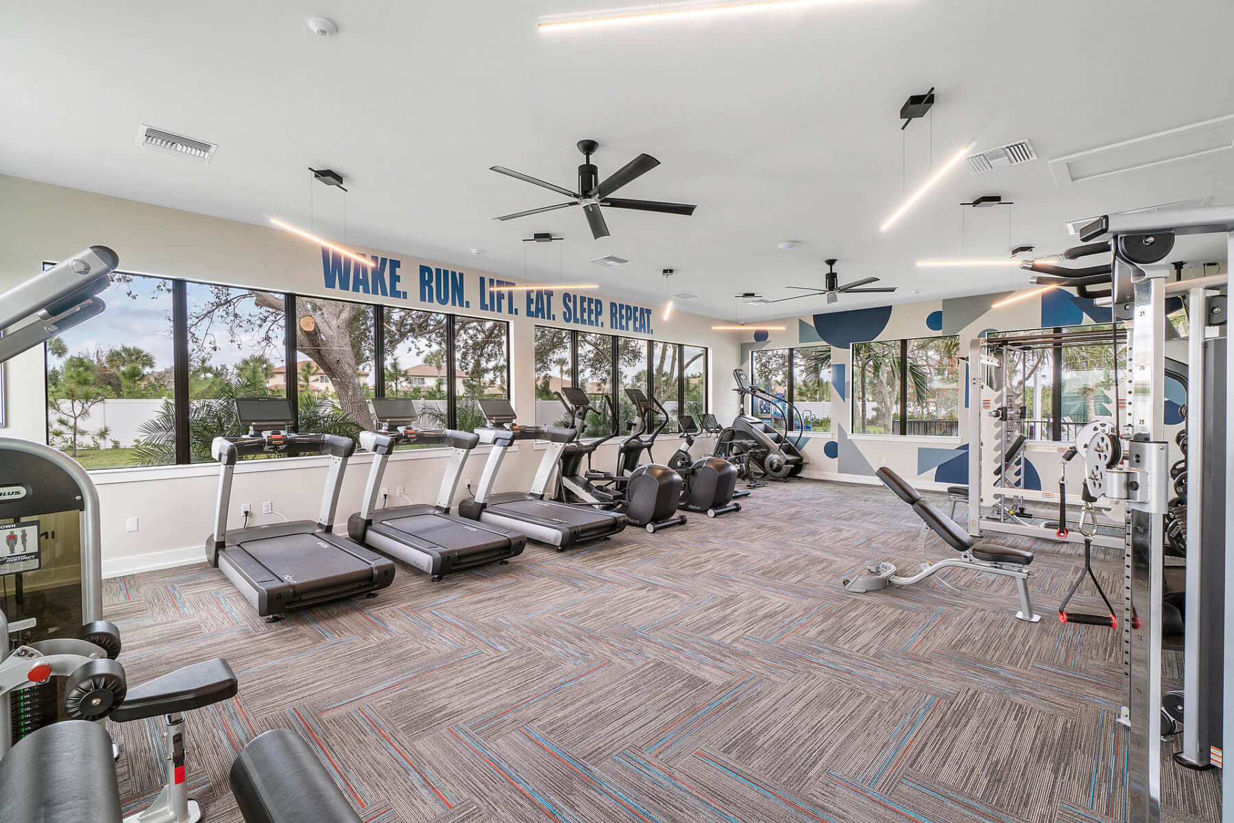 Akel Homes presents a modern gym featuring treadmills, exercise bikes, and weight machines, all set against a serene wooded view through large windows. The walls boast motivational text, and ceiling fans keep the atmosphere fresh and invigorating.