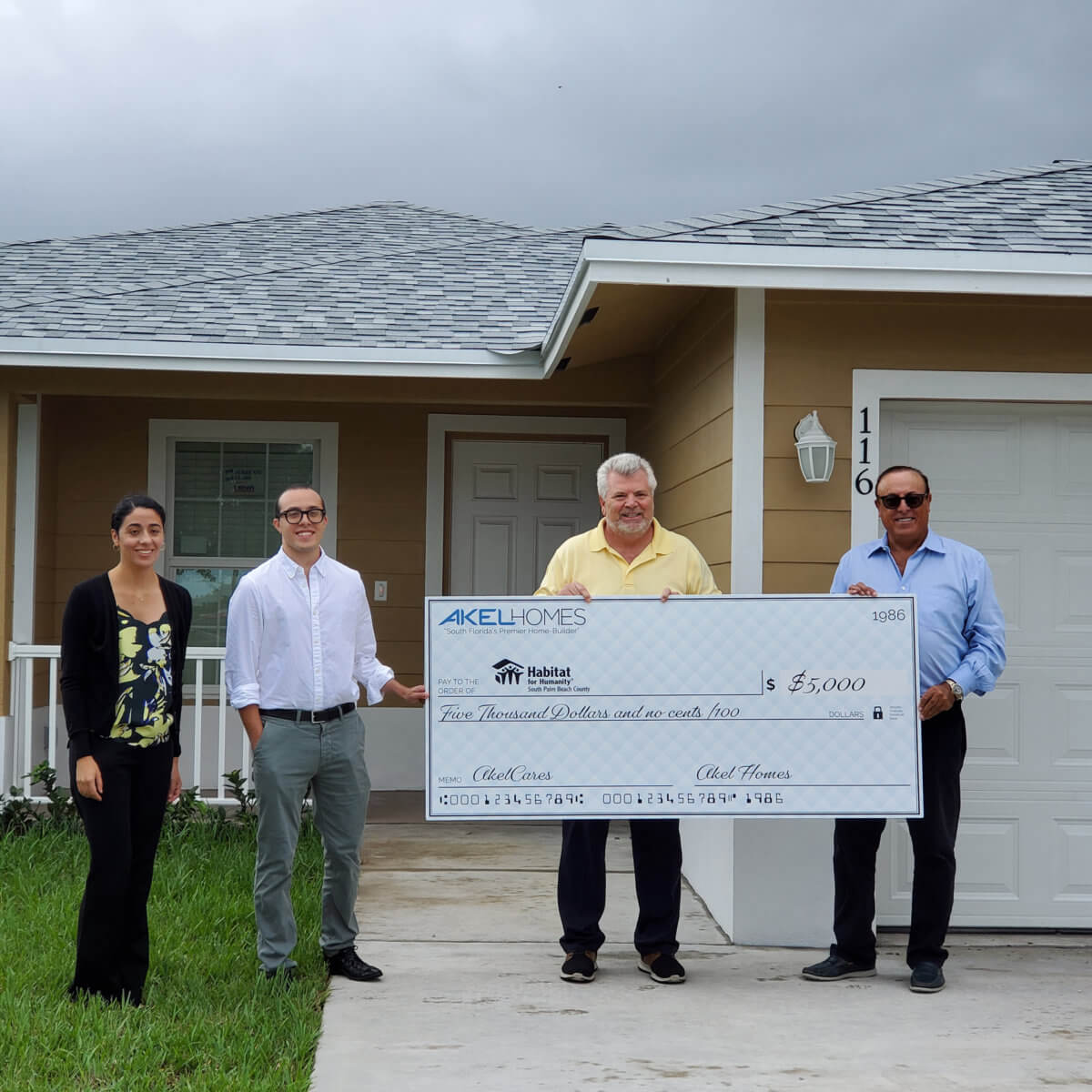 Four people stand in front of a house, proudly holding a large ceremonial check for $5,000 made out to Habitat for Humanity. This generous donation comes courtesy of Akel Homes, highlighting their commitment to supporting community-building initiatives.