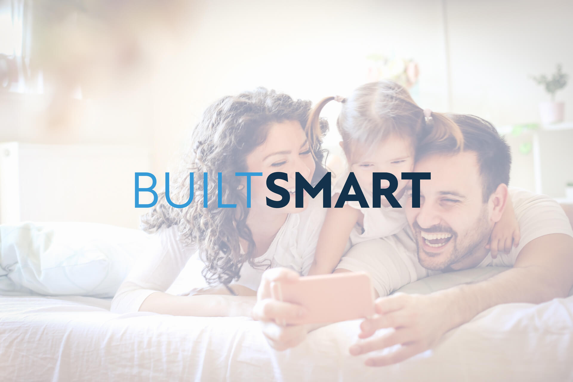 Family lying on a bed, smiling, while looking at a smartphone. Text overlay reads "BUILT SMART with Akel Homes.