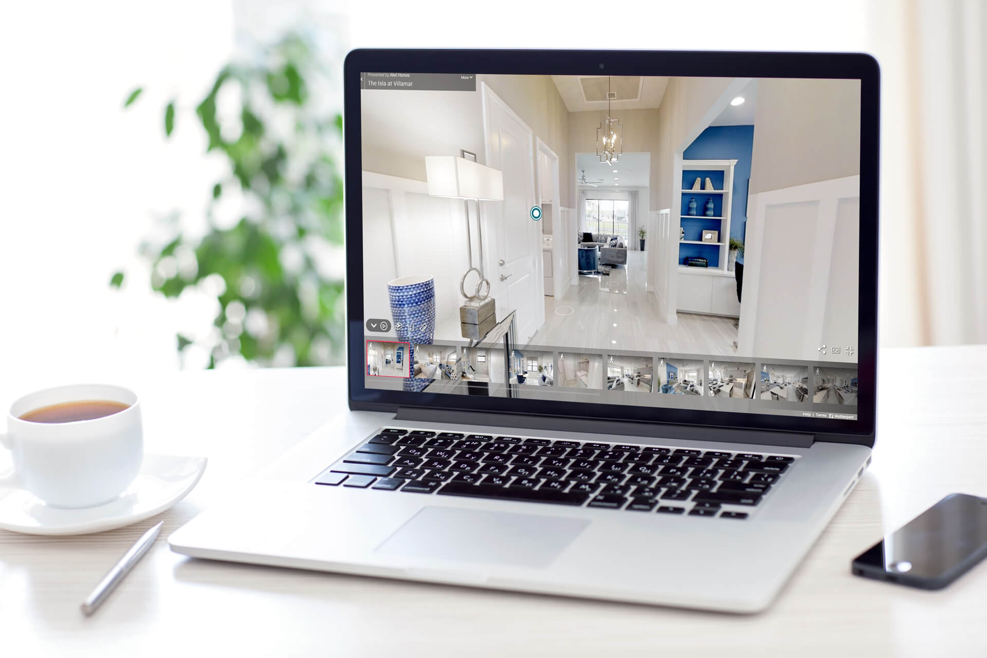 A laptop showcases a virtual tour of a sleek, modern, white-toned interior from Akel Homes. Nearby, a smartphone and a cup of tea rest on the table, completing the cozy scene.