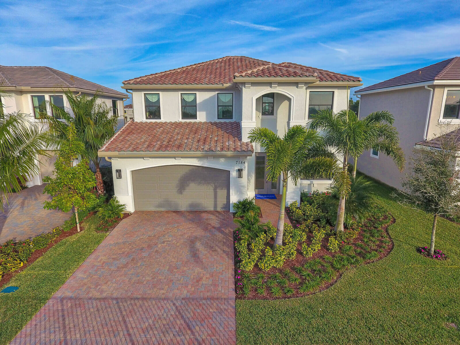 A two-story house by Akel Homes boasts a red-tiled roof, garage, and a landscaped front yard adorned with palm trees and shrubs.