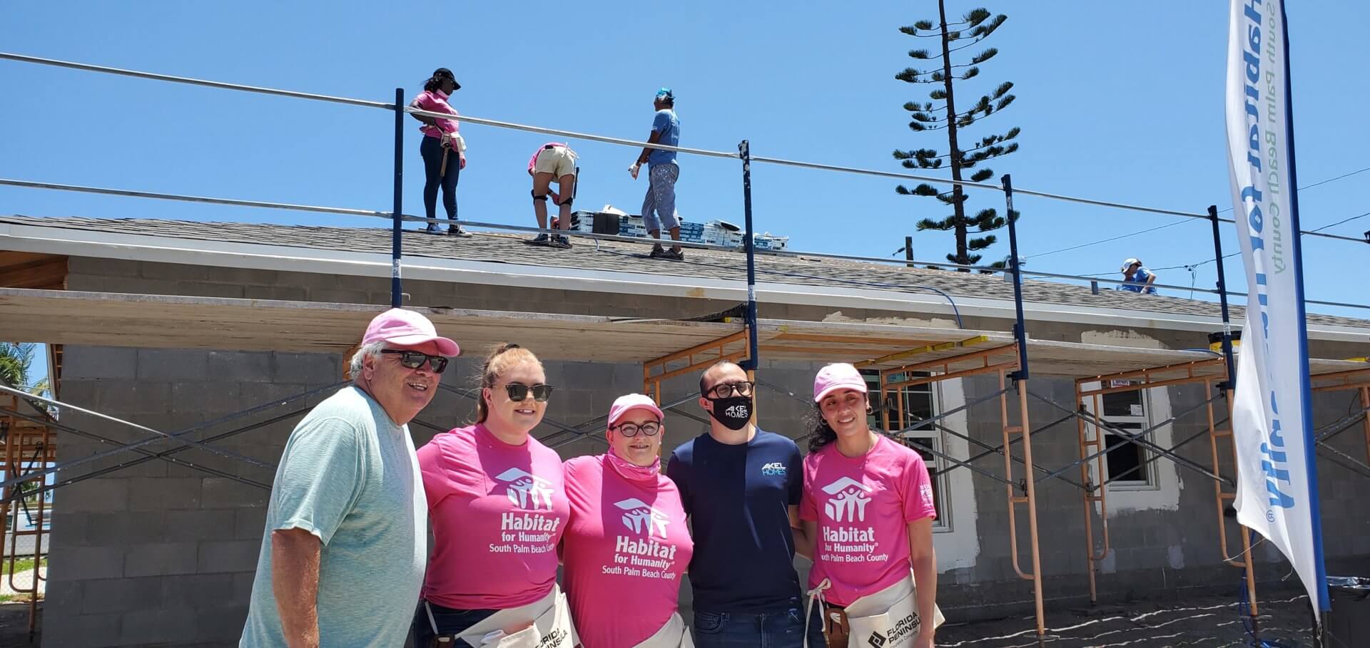 Akel Homes' team, sporting hard hats and pink shirts, gathers proudly in front of a building under construction, with diligent workers seen on the roof.
