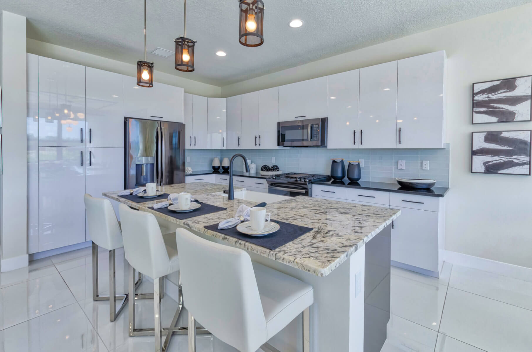 Akel Homes presents a modern kitchen with white cabinets, a granite island, and stainless steel appliances. Three pendant lights illuminate the island, perfectly set with four place settings and sleek white chairs.