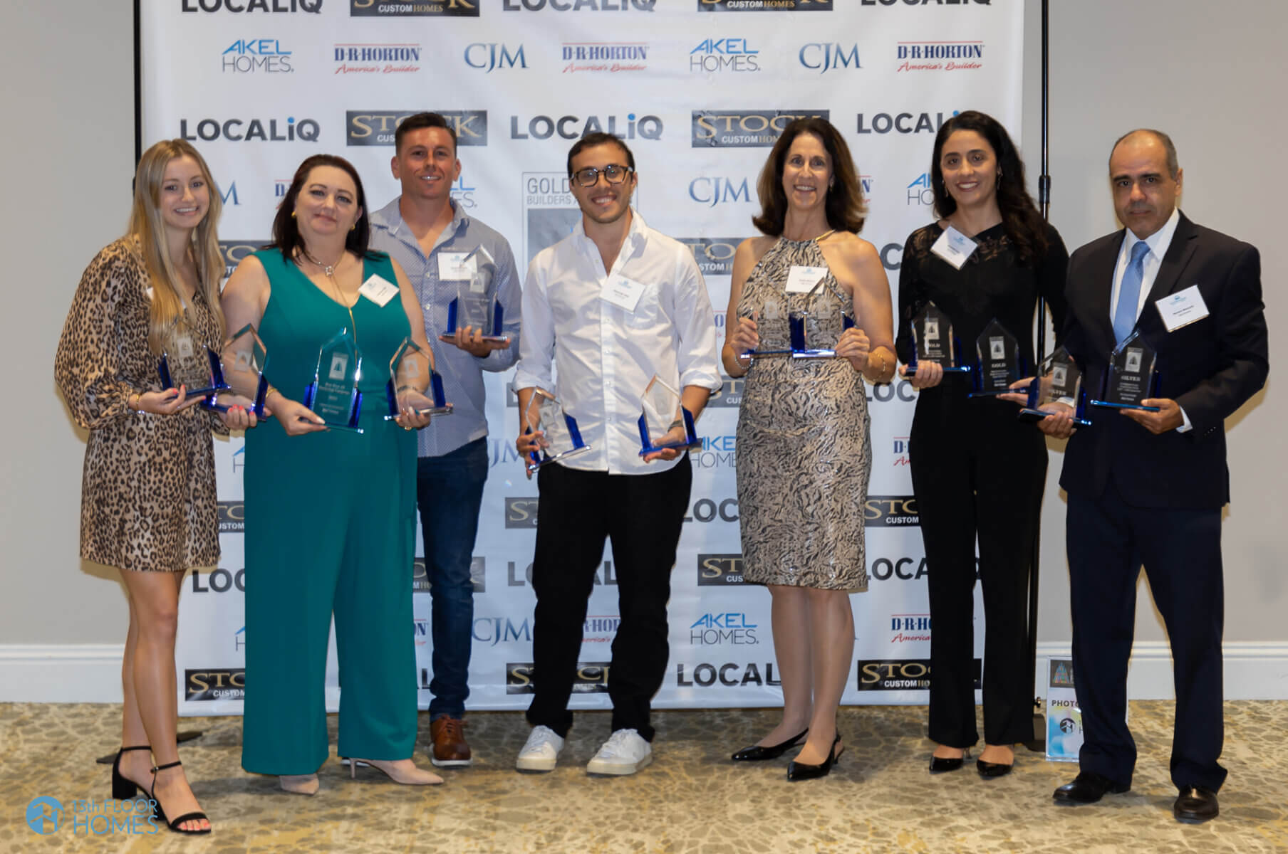A group of eight people stands proudly in front of an Akel Homes banner, each holding a glass award. They are smiling and dressed in business casual and formal attire.