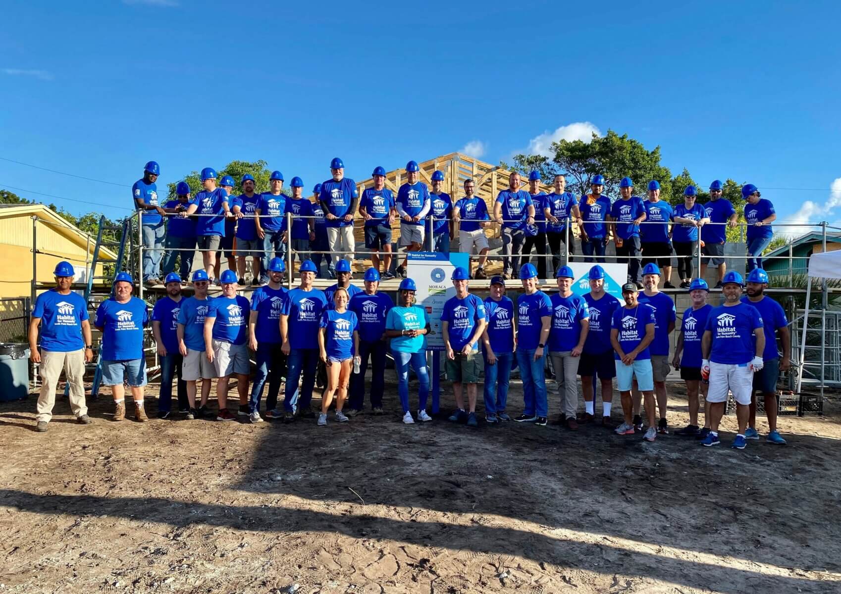 A large group of people in matching blue shirts and hats stand proudly in front of an Akel Homes construction site under a clear blue sky.