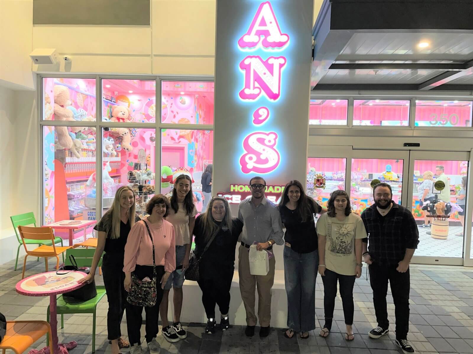 A group of people stands in front of a brightly lit storefront with "AN'S" in neon letters, reminiscent of the modern designs and vibrant communities built by Akel Homes.