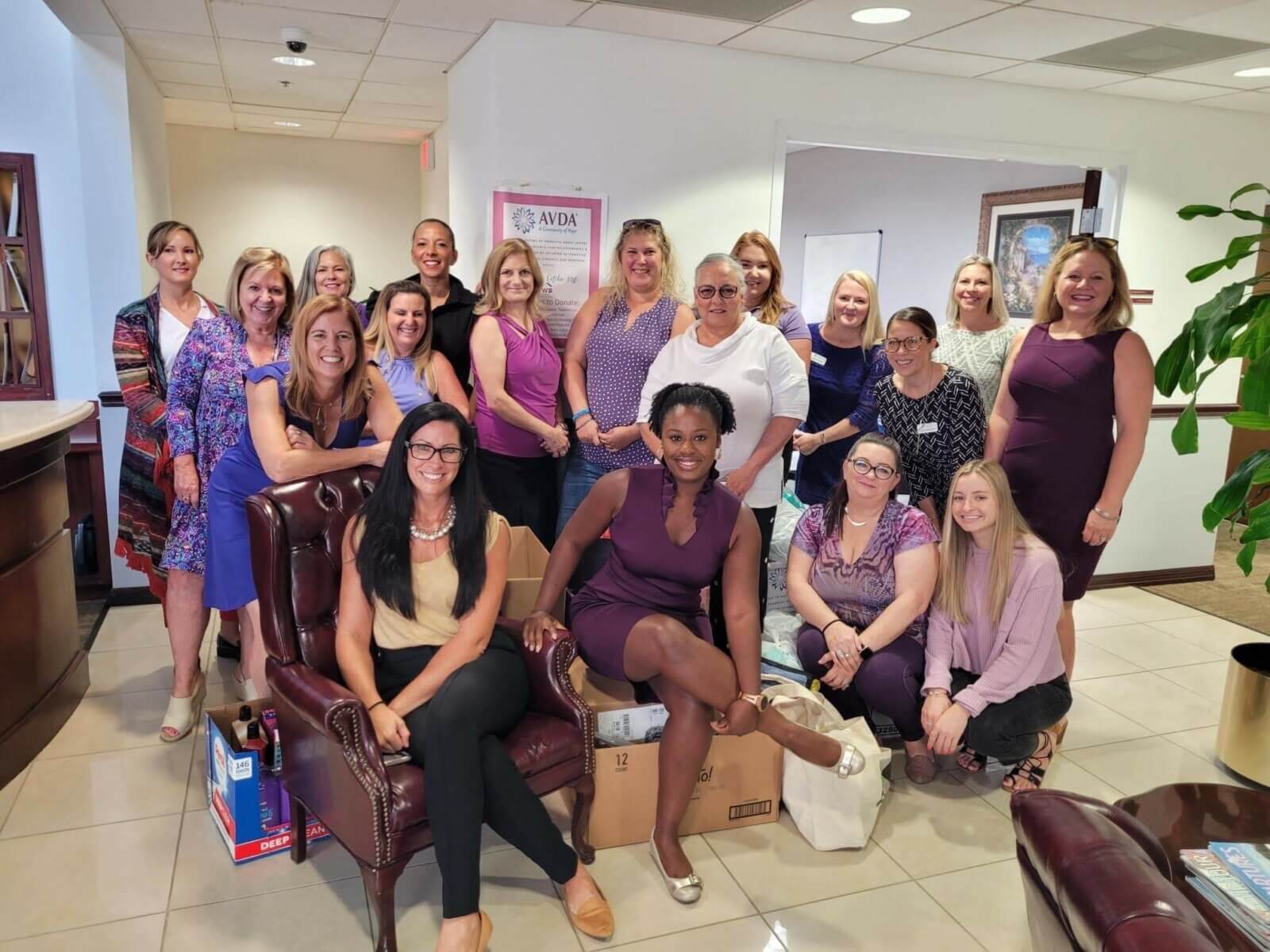 A group of people, mostly women, pose together in an office setting at Akel Homes. Some are seated while others stand behind. They're all smiling, and a plant is visible in the corner.