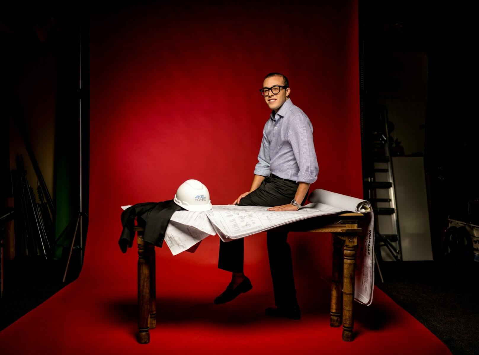 A person in glasses sits on a table covered with Akel Homes blueprints, a hard hat, and a suit jacket, set against a striking red background.