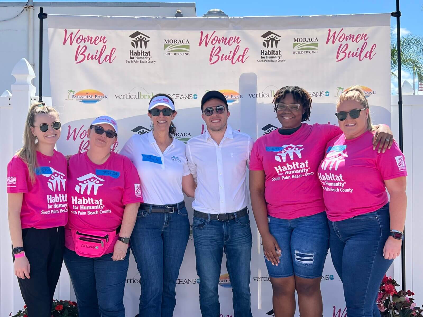 A group of six people, some sporting pink Habitat shirts, stands proudly in front of a Habitat for Humanity banner, showcasing their collaboration with Akel Homes.