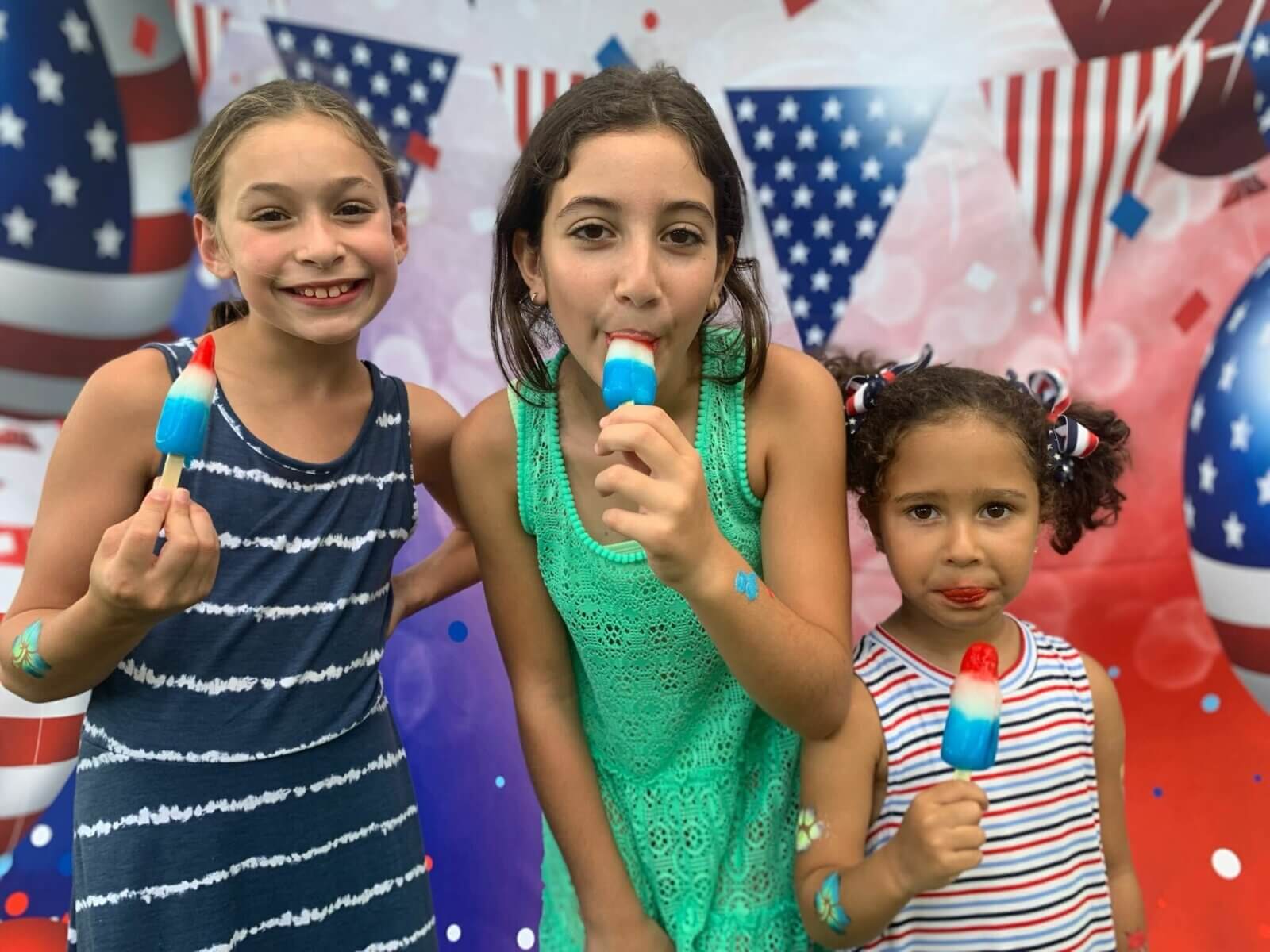 Three children stand joyfully before a patriotic backdrop, holding red, white, and blue popsicles. Their smiles are as bright as the welcoming community of Akel Homes, where every day feels like a celebration of family and freedom.