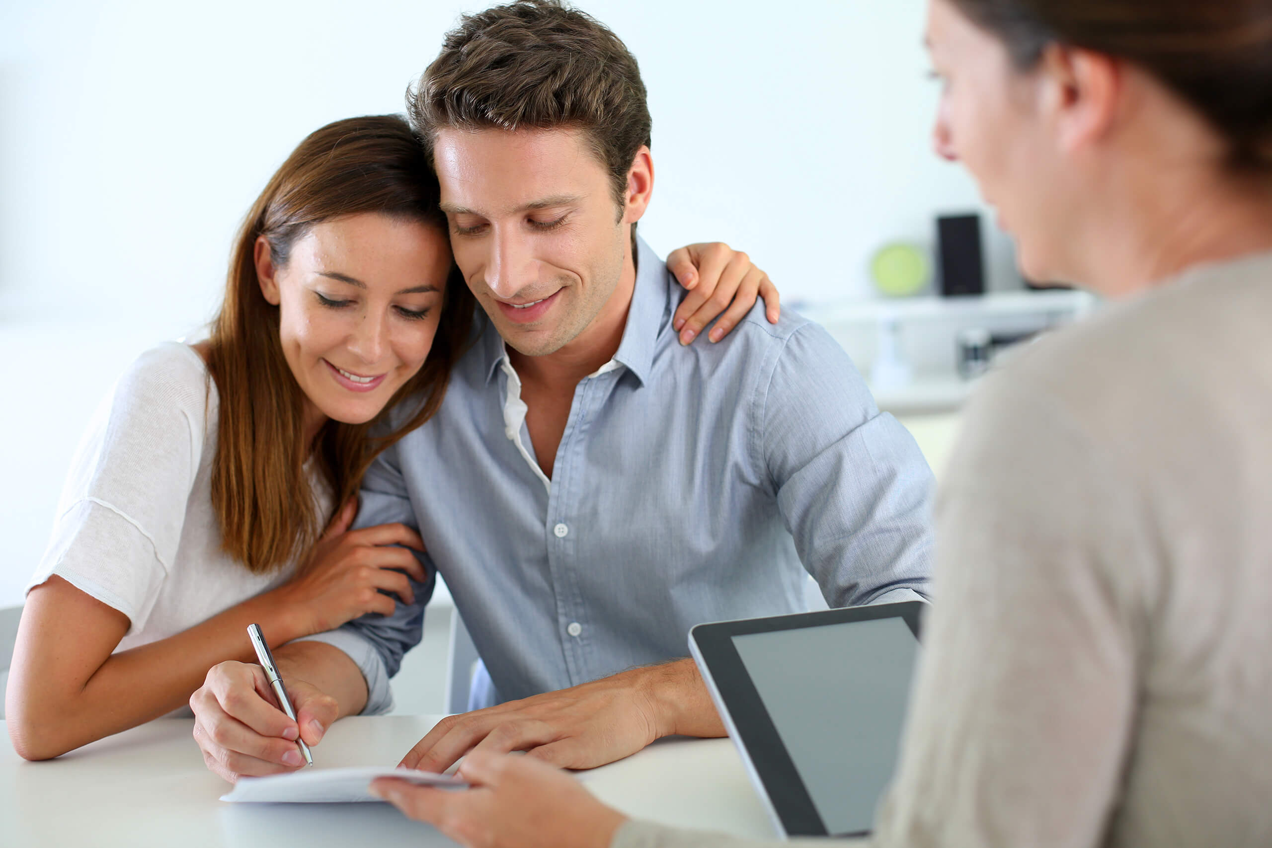 A couple smiles while signing a document at an Akel Homes desk. Another person is seated opposite them, holding a tablet.