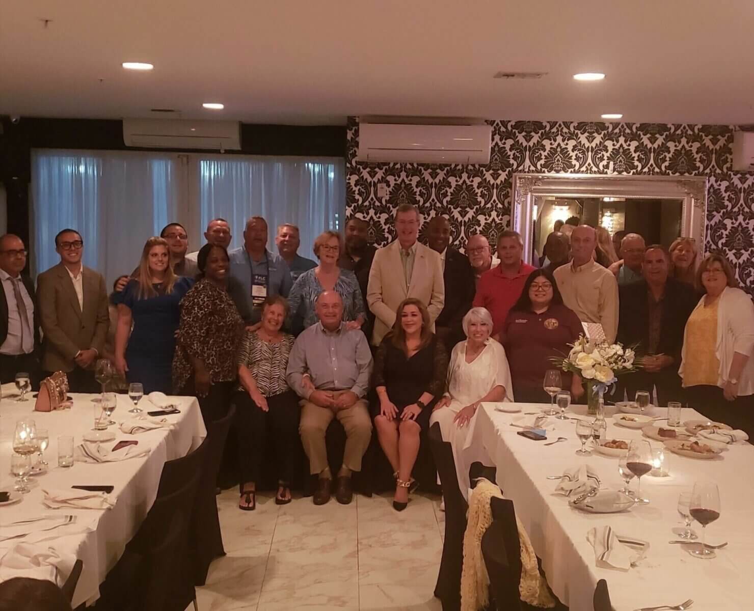 A group of people from Akel Homes are posing together in a dimly lit restaurant. The tables are elegantly set with plates and glasses gleaming softly in the foreground.