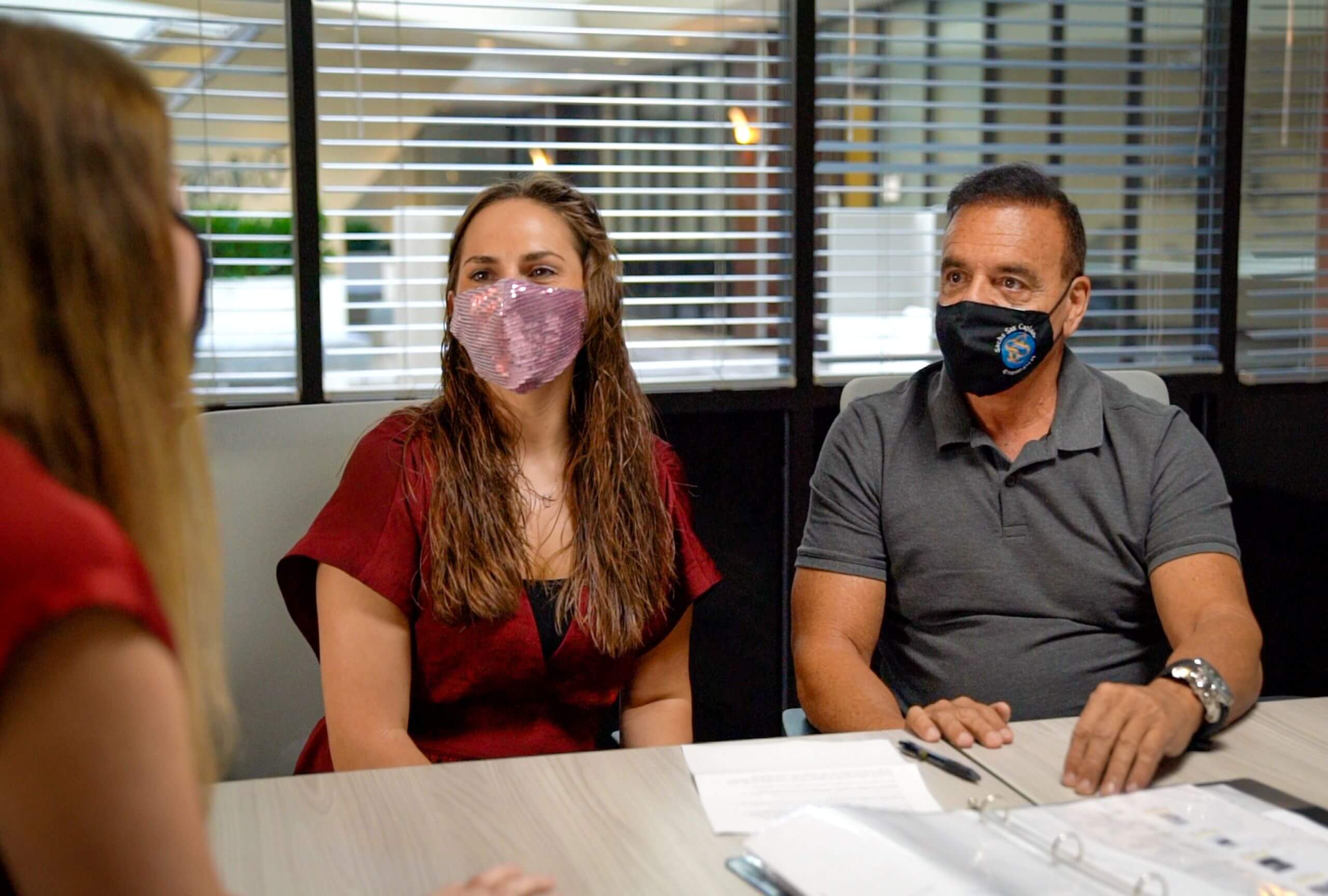 Two people wearing masks sit at a table with documents from Akel Homes, listening intently to a person opposite them in an office setting.