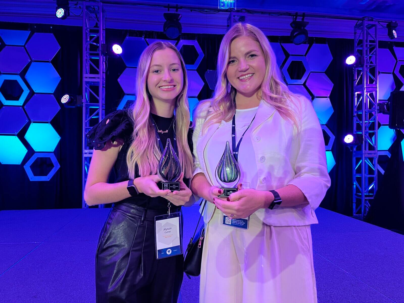 Two women from Akel Homes stand on stage, smiling and holding awards against a vibrant blue and purple hexagonal backdrop.