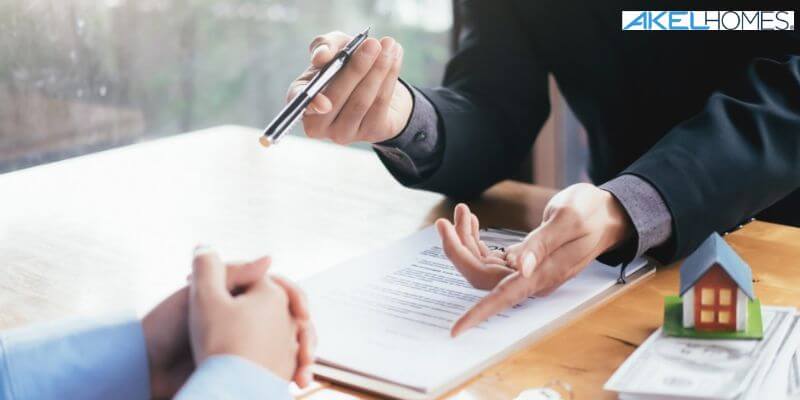 Two people are seated at a table discussing a document. One points to the paper with a pen, while an Akel Homes model sits nearby, adding context to their conversation.