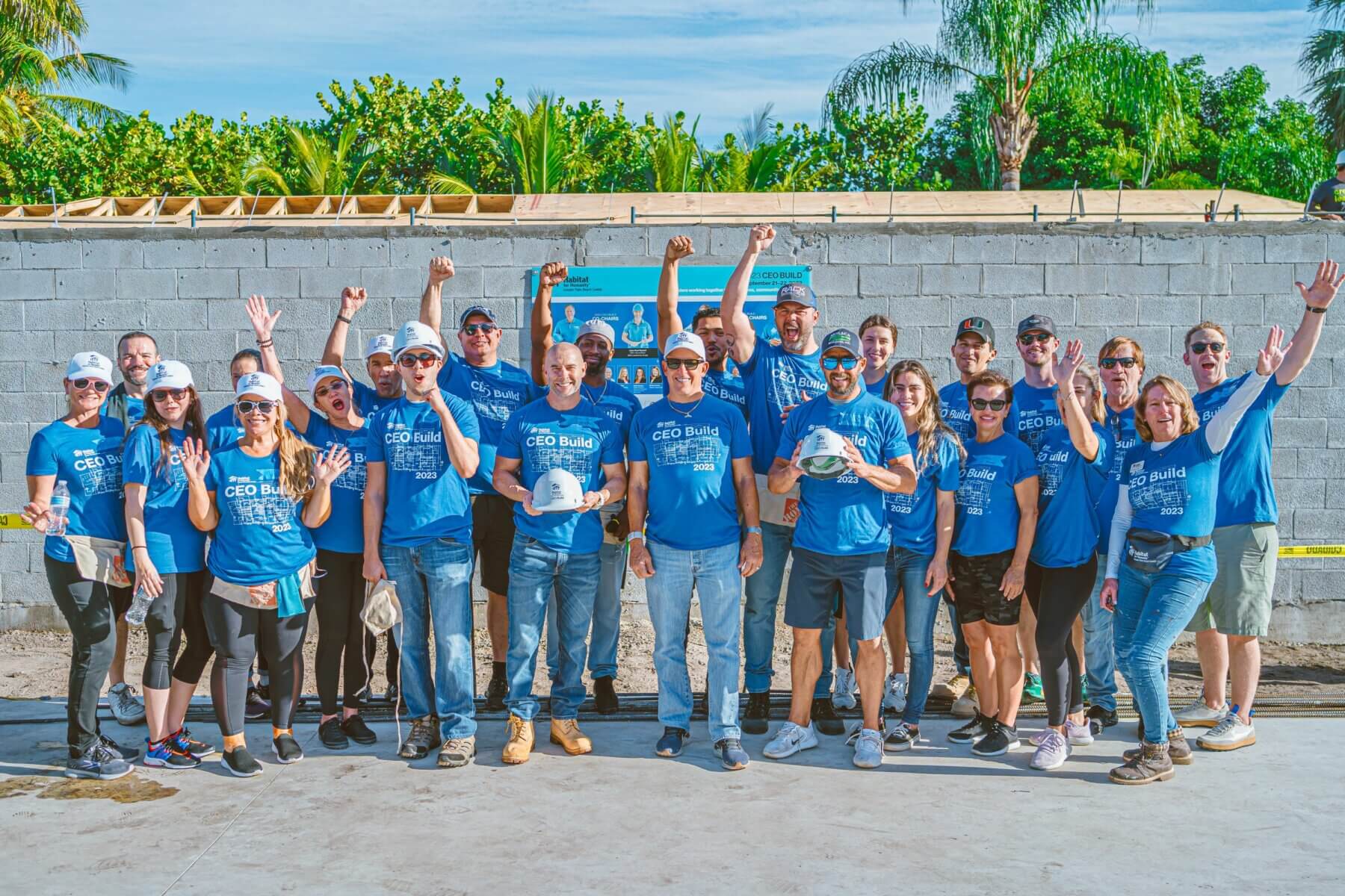 A group of people in blue shirts, proudly representing Akel Homes, pose together in front of a partially constructed wall, smiling and raising their hands. Trees and a clear sky provide the perfect backdrop.