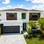This modern two-story house, crafted by Akel Homes, boasts a white facade, dark garage, and large windows. It's embraced by a landscaped yard dotted with palm trees and features a brick driveway under a partly cloudy sky.