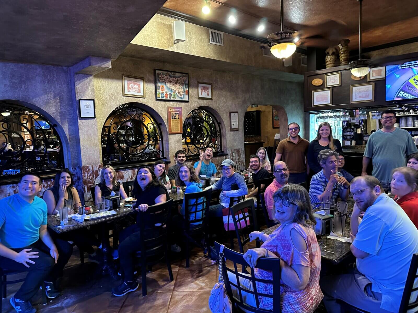 A group of people sit together at tables in a dimly lit restaurant, smiling and facing the camera, as if celebrating the warm sense of community that Akel Homes fosters.