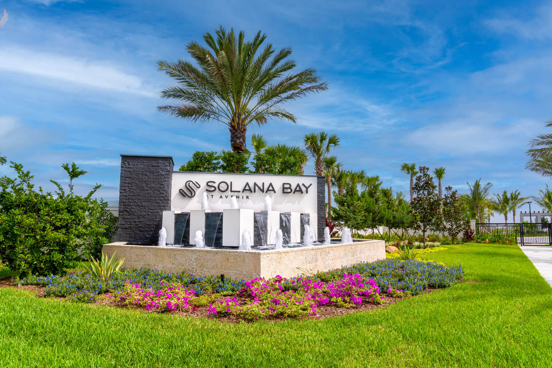 The entrance sign to Solana Bay by Avenir, crafted by Akel Homes, showcases vibrant landscaping with palm trees, colorful flower beds, and five elegant water fountains sparkling under the sunny sky.