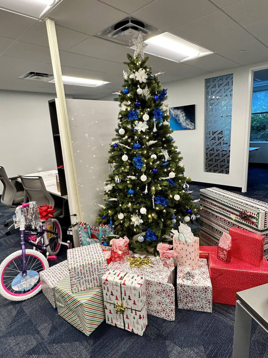 The Akel Homes office Christmas tree stands adorned with blue and white ornaments, creating a festive focal point surrounded by wrapped gifts and a children's bicycle.