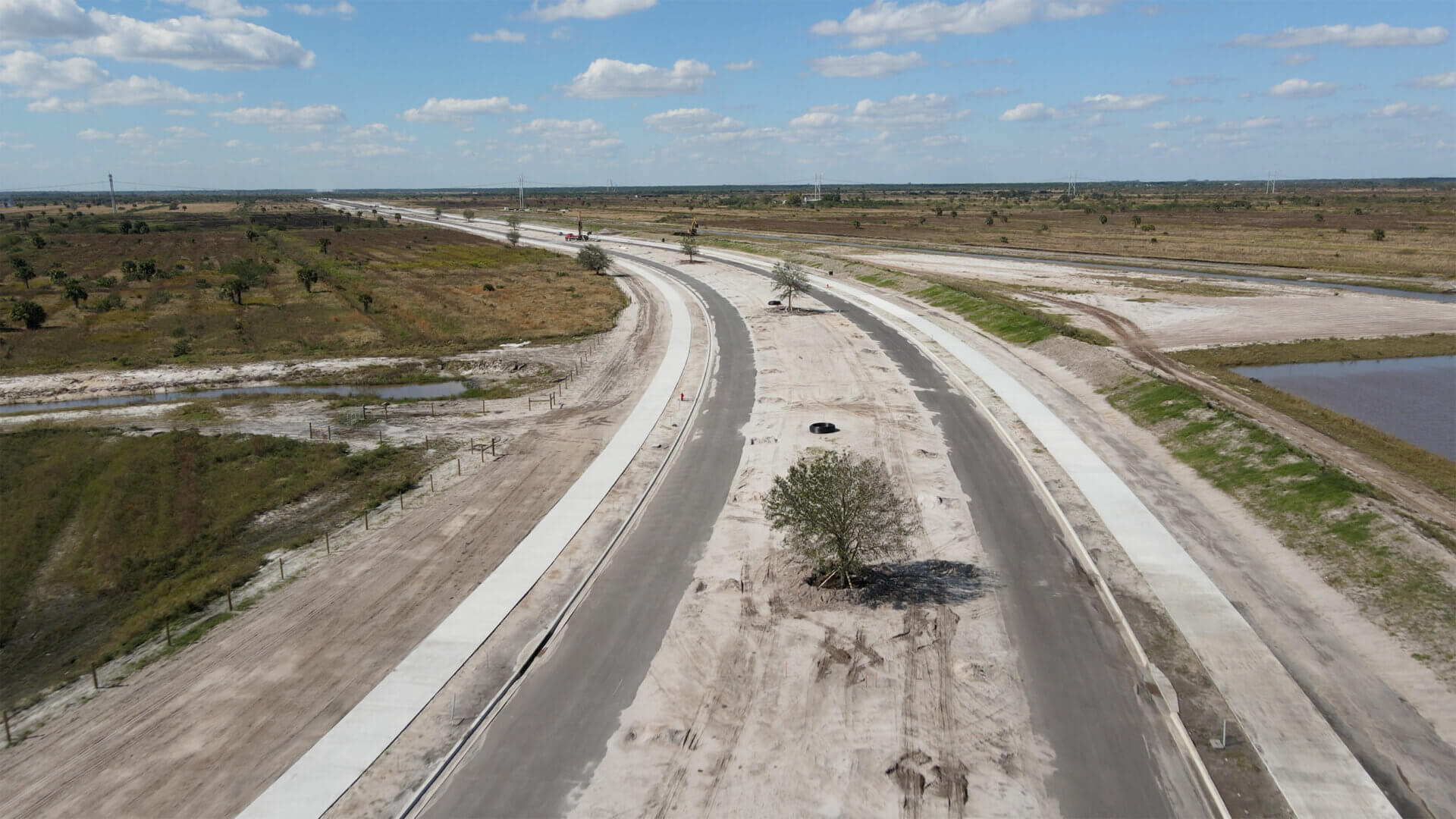 Aerial view of a newly paved, deserted road with surrounding open land under a blue sky with scattered clouds, highlighting the pristine landscapes often developed by Akel Homes.