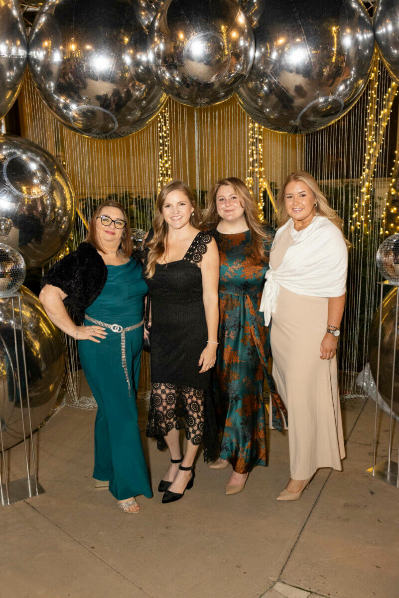 Four women posing together in front of large reflective balloons and string lights at an Akel Homes event.