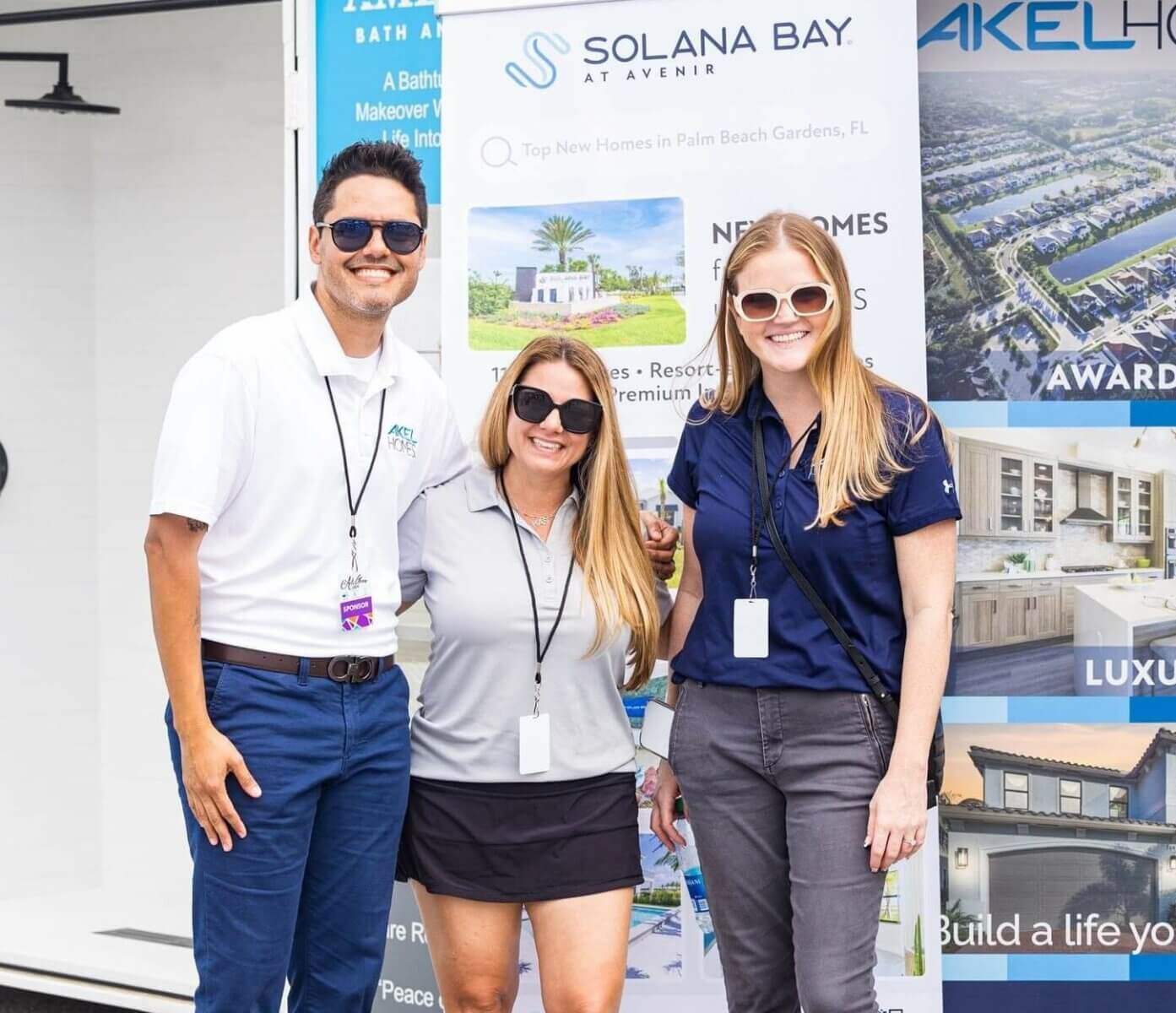 Three people, each wearing sunglasses and name badges, smile warmly in front of promotional banners for Solana Bay. Their presence exudes the welcoming and innovative spirit synonymous with Akel Homes.