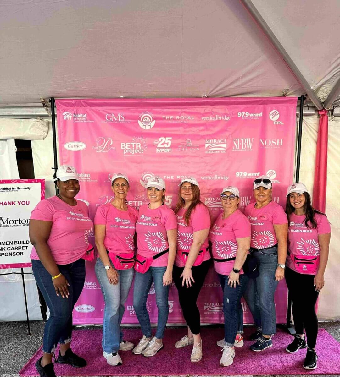 A group of seven people in pink shirts and caps stand proudly in front of a pink Akel Homes sponsor backdrop at an outdoor event.