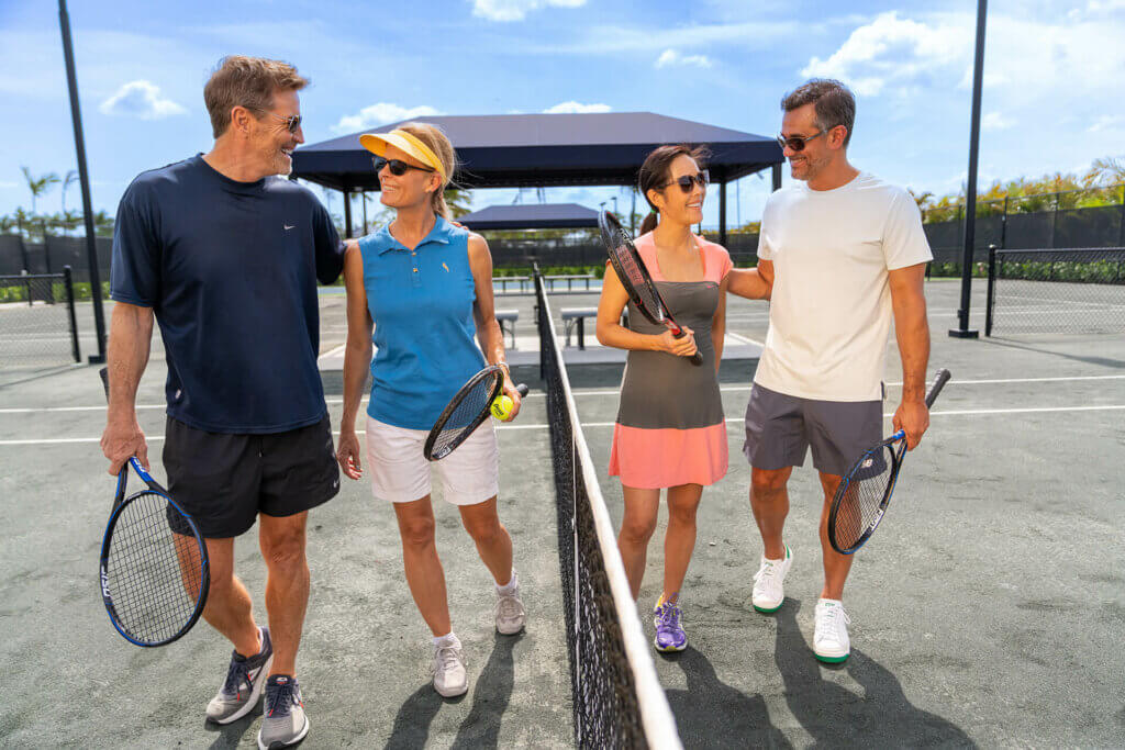 Four adults with tennis rackets, wearing Akel Homes attire, stroll onto a tennis court, engaged in lively conversation under a clear blue sky.