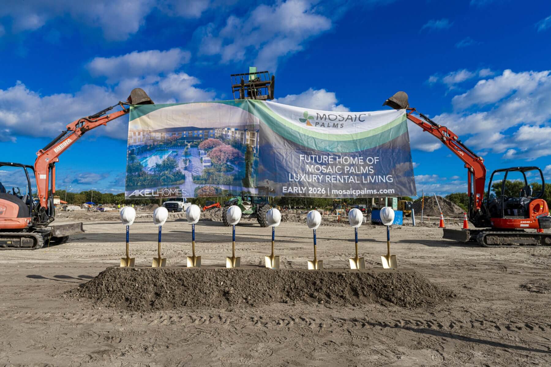 At the construction site where dreams take root, a banner proudly announces the "Future Home of Mosaic Palms Luxury Rental Living, Early 2026." In the foreground, eight hard hats rest on shovels signifying groundbreaking progress, as two excavators stand ready behind.