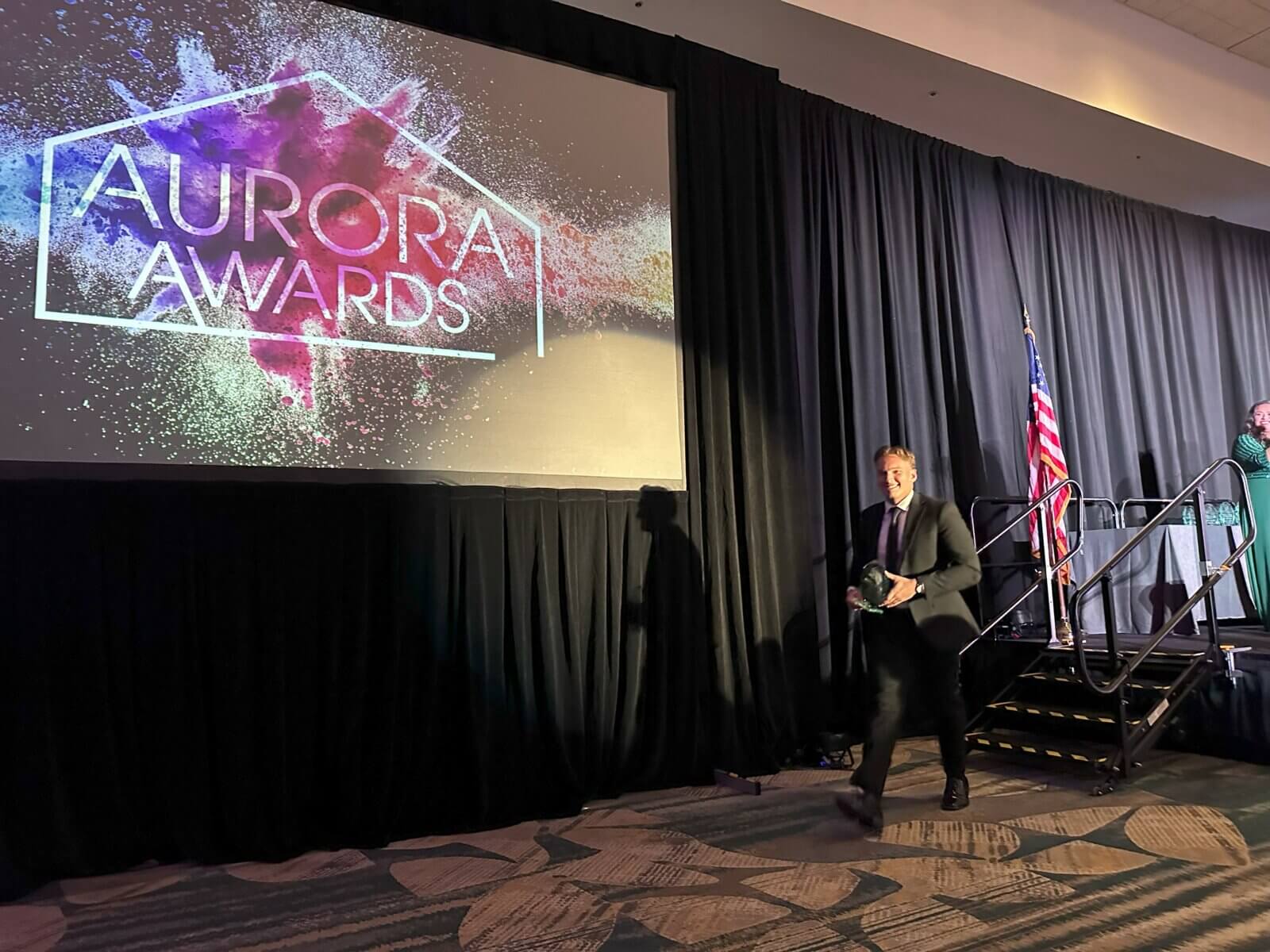 A person walks on stage during the Aurora Awards ceremony, with a colorful screen displaying the event name. An American flag is visible in the background, subtly echoing the pride of Akel Homes in their dedication to excellence.