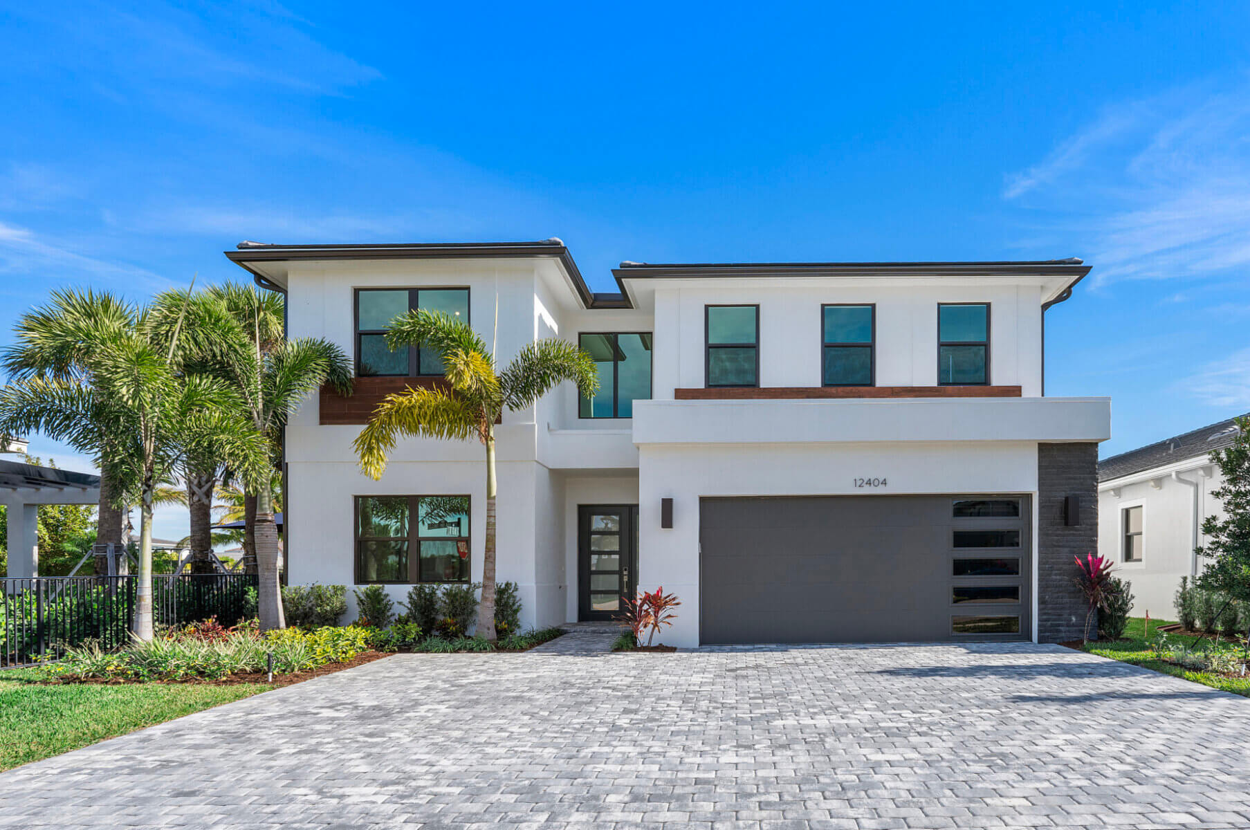 Discover the elegance of this modern two-story Solana Bay home by Akel Homes, featuring a flat roof and expansive windows. This quick move-in home includes a two-car garage, paved driveway, and a lush front yard adorned with palm trees and vibrant grass.