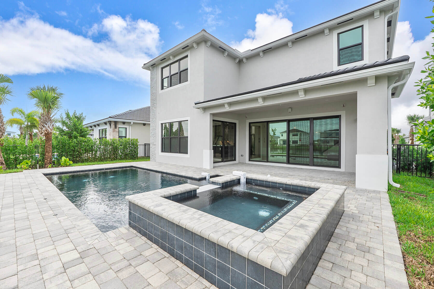 Modern two-story house with large windows, a gray exterior, and a backyard featuring a rectangular swimming pool surrounded by stone tiles and green landscaping.