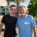 Two men stand outdoors smiling at the camera, with a food truck and palm trees in the background. One man wears a black shirt; the other has a name tag and a blue patterned shirt, near the vibrant SB Sculpture.