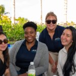 Four women sit at an outdoor table with drinks and food, smiling at the camera. Palm trees, greenery, and a glimpse of the SB Sculpture create a lively backdrop.