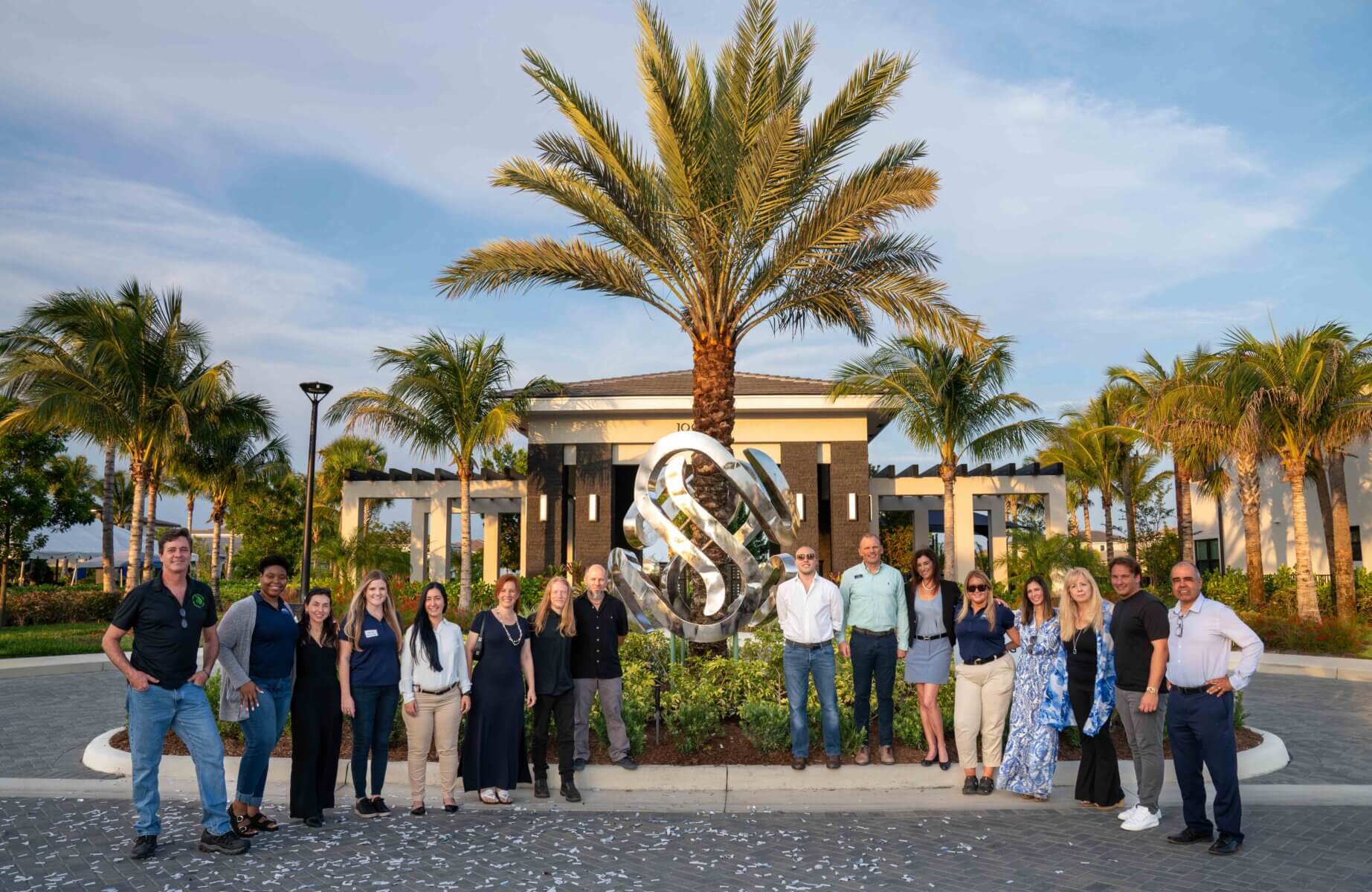 A group of people stands in front of a building and palm trees, posing around a large metallic sculpture with an interlocking design at Solana Bay by Akel Homes.