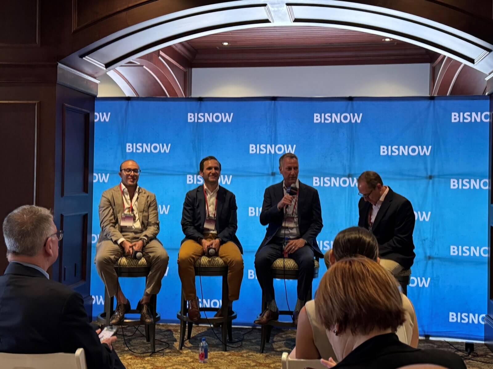 Four men in business attire sit on stools on a panel stage with a blue BISNOW backdrop, discussing the State of the Market and trends impacting Akel Homes and the Treasure Coast, while speaking to an audience at an indoor event.