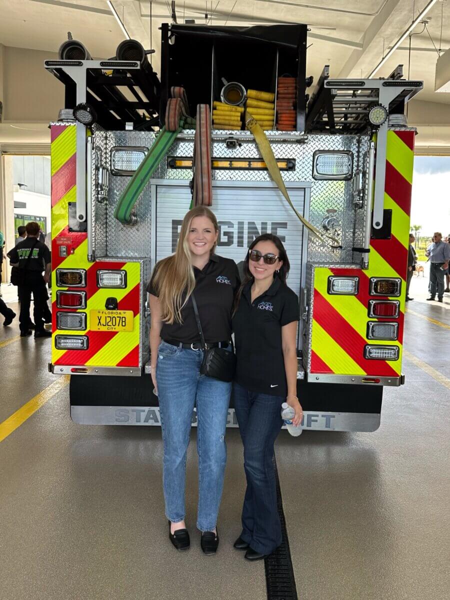 Two women wearing black shirts and jeans stand smiling in front of a fire truck inside a station.