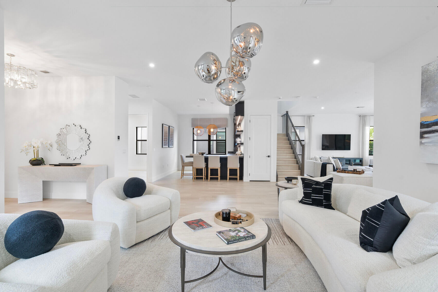 Modern open-plan living room with white sofas, round coffee table, black accent pillows, and metallic pendant lights; kitchen and dining area visible in the background—an elegant showcase of luxury new construction homes.