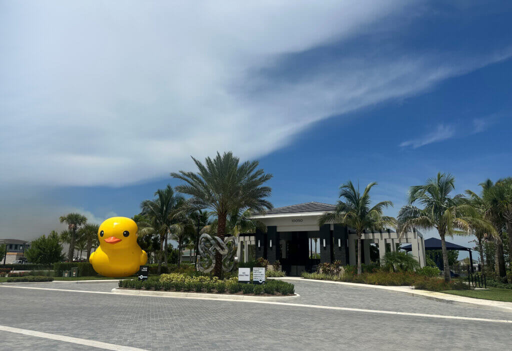 A large inflatable yellow duck, part of the Luckiest Duck Program, is displayed near the entrance of Solana Bay's modern building surrounded by palm trees and landscaping under a partly cloudy sky.