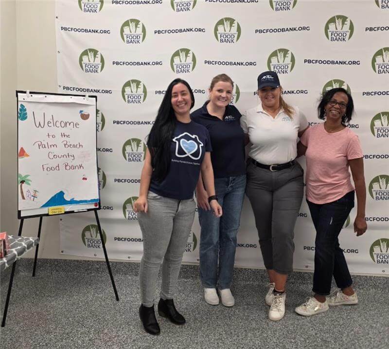 Four women stand smiling in front of a Palm Beach County Food Bank banner next to a welcome sign inside the facility, proudly representing Akel Homes Volunteers.