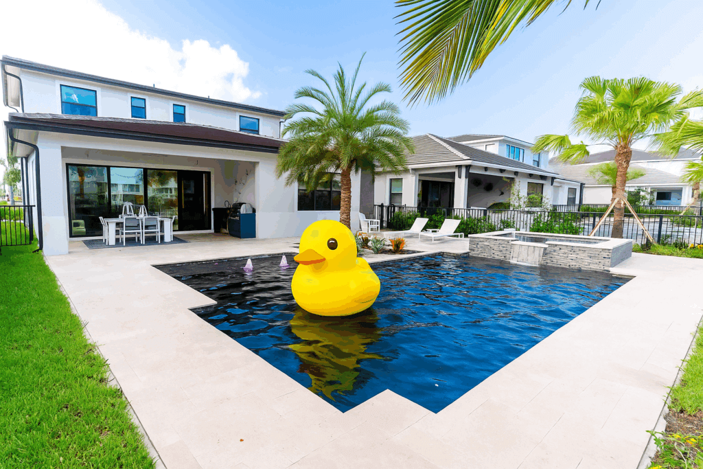 Modern backyard with a swimming pool featuring a large inflatable yellow duck, patio furniture, palm trees, and a contemporary house in the background—perfect for your next Facebook Live Event.